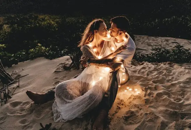 man and woman looking each other sitting on white sand during sunset